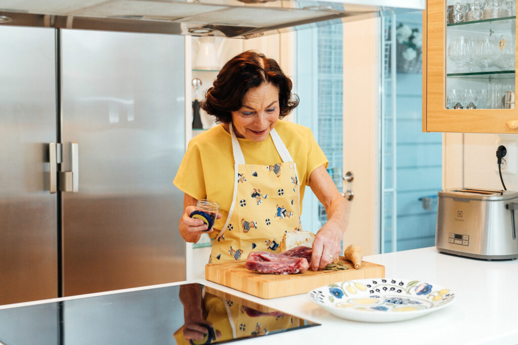 Nainen keltaisessa t-paidassa ja essussa tekee liharuokaa keittiössä.